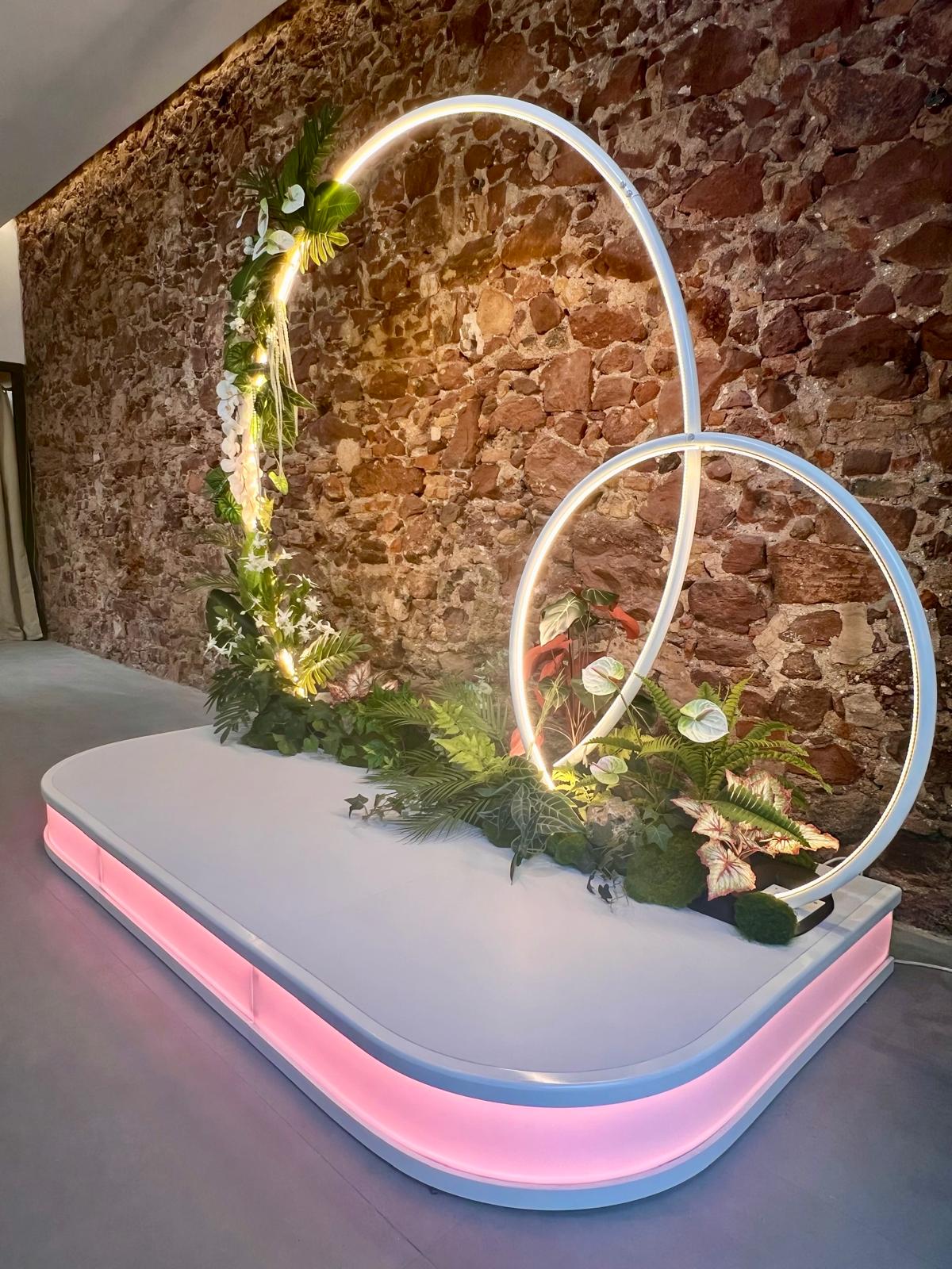Photocall escultórico con aros de luz LED entrelazados y decoración floral sobre plataforma iluminada